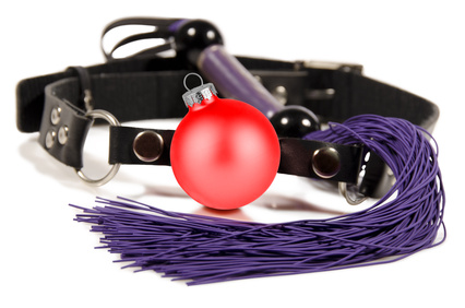 Arrangement aus BDSM-Zubehör: ein Lederhalsband und ein kurzer, lilafarbener Flogger - dekoriert mit einer roten Weihnachtskugel vor weißem Hintergrund.